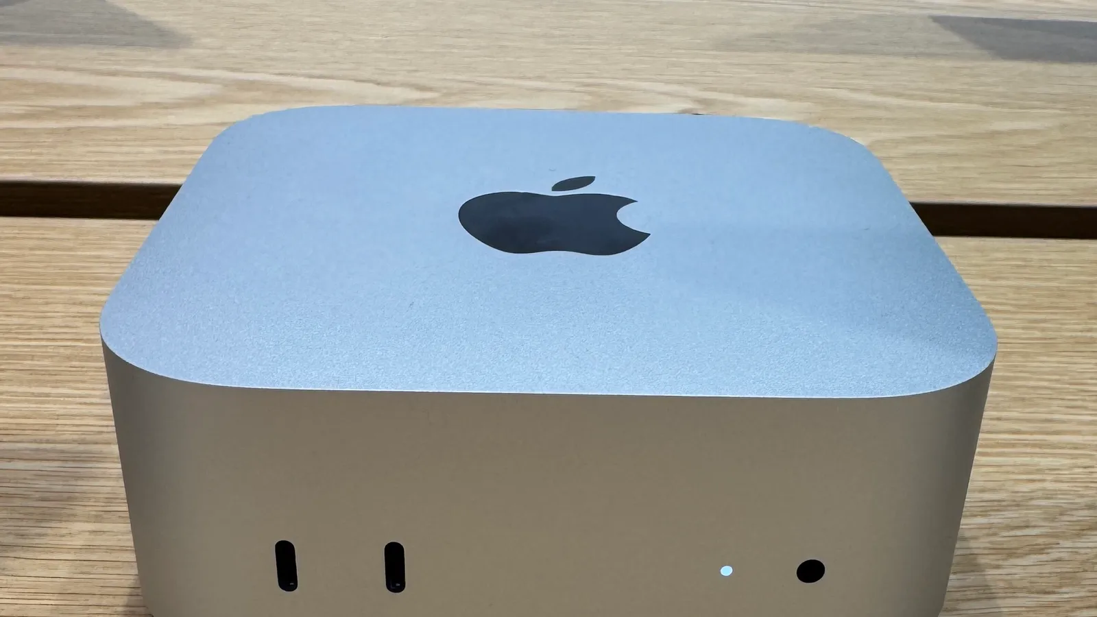 A silver Apple Mac mini with its compact square case, front ports visible, on a wooden table.
